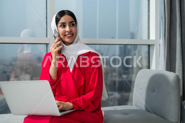 النظر إلى شيء ما أثناء إجراء مكالمة هاتفية عبر الجوال، الجلوس في لوبي إحدى الفنادق الراقية، استخدام الأجهزة التكنولوجية الحديثة والمتطورة، صورة مقربة لإمرأة عربية خليجية سعودية مبتسمة ترتدي الحجاب تعمل على اللابتوب وتقوم بالدردشة والتحدث على الهاتف المحمول