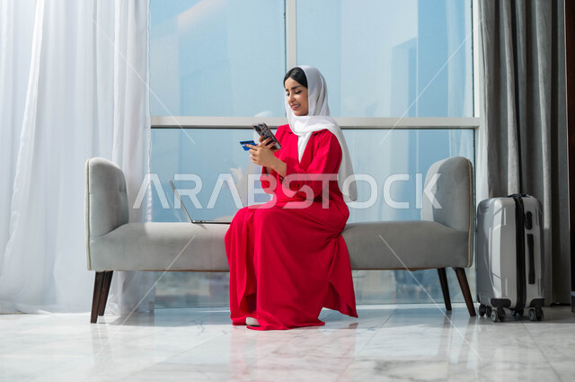 استخدام التقنيات الحديثة والمتطورة، إمرأة عربية خليجية سعودية مبتسمة ترتدي الحجاب تمسك بيدها الهاتف المحمول وبطاقة الائتمان البنكية تجلس بجانب حقيبة السفر، إمكانية حجز تذاكر الطيران والإقامة الفندقية عبر الأونلاين، الشراء والدفع من خلال برامج وتطبيقات الجوال