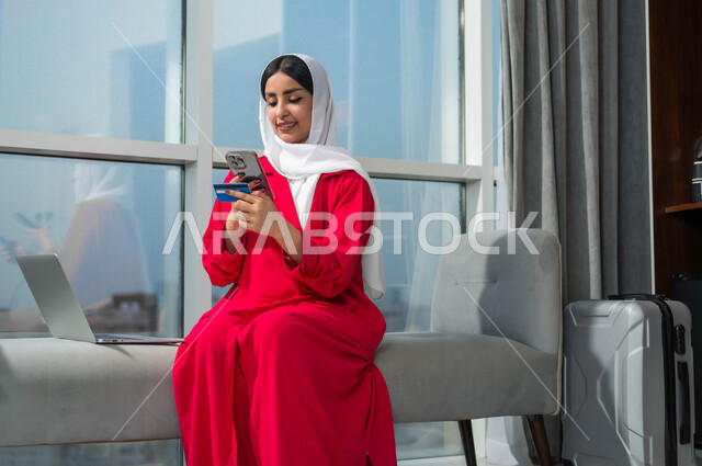 إمكانية حجز تذاكر الطيران والإقامة الفندقية عبر الأونلاين، الشراء والدفع من خلال برامج وتطبيقات الجوال، استخدام التقنيات الحديثة والمتطورة، إمرأة عربية خليجية سعودية مبتسمة ترتدي الحجاب تمسك بيدها الهاتف المحمول وبطاقة الائتمان البنكية تجلس بجانب حقيبة السفر