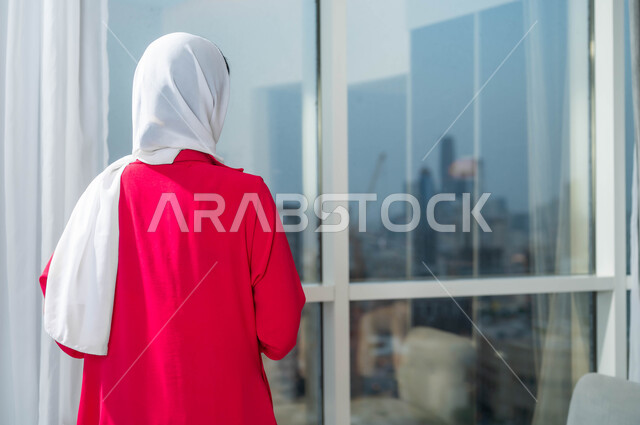 التأمل وشرود الذهن، التفكير في شيء ما، صورة مقربة من الخلف لإمرأة عربية خليجية سعودية ترتدي الحجاب تقف امام واجهه زجاجية كبيرة في أحد الأبراج الراقية، الاستمتاع بالنظر الى العالم الخارجي من خلال النافذة