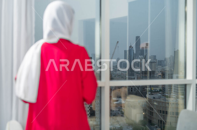 التفكير في شيء ما، صورة مقربة من الخلف لإمرأة عربية خليجية سعودية ترتدي الحجاب تقف امام واجهه زجاجية كبيرة في أحد الأبراج الراقية، الاستمتاع بالنظر الى العالم الخارجي من خلال النافذة، التأمل وشرود الذهن