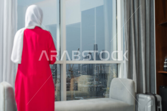 الاستمتاع بالنظر الى العالم الخارجي من خلال النافذة، التأمل وشرود الذهن، التفكير في شيء ما، صورة مقربة من الخلف لإمرأة عربية خليجية سعودية ترتدي الحجاب تقف امام واجهه زجاجية كبيرة في أحد الأبراج الراقية