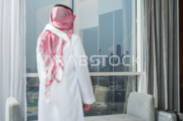 التفكير في شيء ما، صورة مقربة من الخلف لرجل عربي خليجي سعودي يرتدي الشماغ والثوب السعودي التقليدي يقف امام واجهه زجاجية كبيرة في أحد الأبراج الراقية، الاستمتاع بالنظر الى العالم الخارجي من خلال النافذة، التأمل وشرود الذهن
