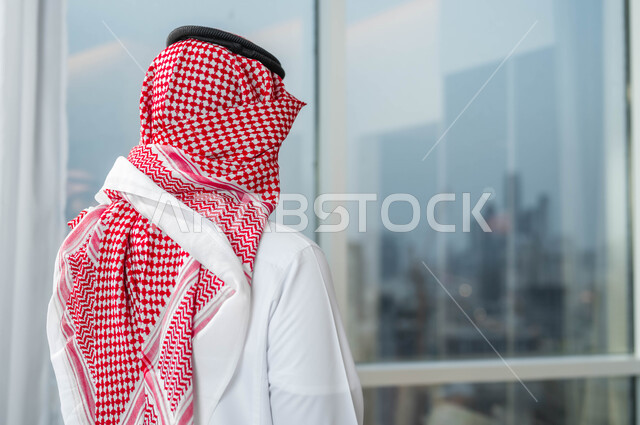 الاستمتاع بالنظر الى العالم الخارجي من خلال النافذة، التأمل وشرود الذهن، التفكير في شيء ما، صورة مقربة من الخلف لرجل عربي خليجي سعودي يرتدي الشماغ والثوب السعودي التقليدي يقف امام واجهه زجاجية كبيرة في أحد الأبراج الراقية