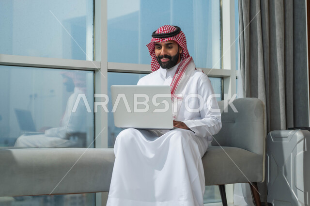 الجلوس في إحدى الفنادق الراقية، العمل عبر الحاسب المحمول، استخدام التقنيات الحديثة والمتطورة، رجل عربي خليجي سعودي يرتدي الشماغ والثوب التقليدي يجلس في لوبي في فندق بجانبه حقيبة السفر ويستخدم اللابتوب