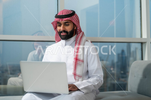 إيماءات الاندماج والتركيز أثناء العمل عبر الحاسب المحمول، الجلوس في إحدى الفنادق الراقية، استخدام الأجهزة التكنولوجية الحديثة والمتطورة، رجل عربي خليجي سعودي يرتدي الشماغ والثوب التقليدي يجلس في لوبي فندق يستخدم اللابتوب