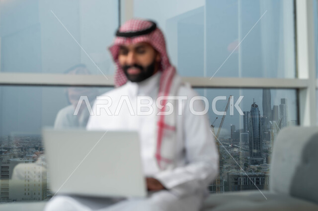 تعابير الاندماج والتركيز أثناء العمل عبر الحاسب المحمول، الجلوس في إحدى الفنادق الراقية، استخدام الأجهزة التكنولوجية الحديثة والمتطورة، رجل عربي خليجي سعودي يرتدي الشماغ والثوب التقليدي يجلس في لوبي فندق يستخدم اللابتوب