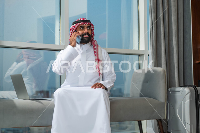 Sitting in an upscale hotel, talking on a mobile phone, using modern and advanced technologies, a Saudi Gulf Arab man wearing a shemagh and a traditional thobe is sitting in the lobby of a hotel with his suitcase next to him and making a phone call.