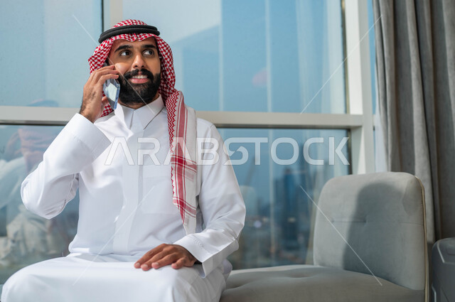 إجراء مكالمة هاتفية عبر الجوال، الجلوس في إحدى الفنادق الراقية، استخدام الأجهزة التكنولوجية الحديثة والمتطورة، رجل عربي خليجي سعودي يرتدي الشماغ والثوب التقليدي يجلس في بهو فندق راقي يقوم بالتحدث عبر الهاتف المحمول وينظر إلى شيء ما