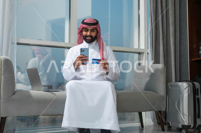 إمكانية حجز تذاكر الطيران والفنادق عبر الأونلاين، الشراء والدفع من خلال برامج وتطبيقات الجوال،استخدام التقنيات الحديثة والمتطورة، رجل عربي خليجي سعودي مبتسم يرتدي الشماغ والثوب التقليدي يمسك بيده الهاتف المحمول وبطاقة الائتمان البنكية
