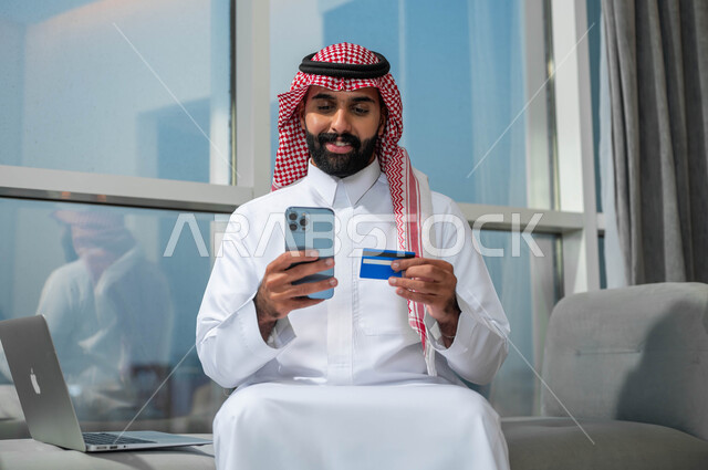 Purchasing and paying through mobile programs and applications, the ability to pay bills with an electronic card, gestures of happiness and joy using modern technologies, a smiling Saudi Arabian Gulf man wearing a shemagh and traditional thobe holding a mobile phone and a bank credit card in his hand.