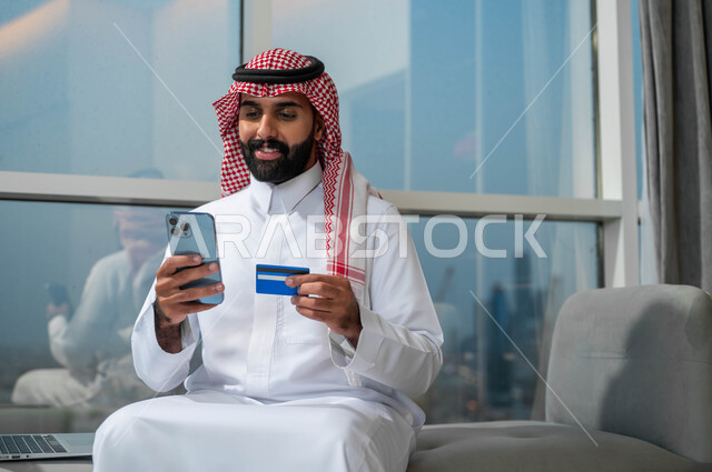 The possibility of paying bills with an electronic card, gestures of happiness and joy using modern technologies, purchasing and paying through mobile programs and applications, a smiling Saudi Arabian Gulf man wearing a shemagh and traditional thobe, holding a mobile phone and a bank credit card in his hand.