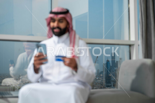 Gestures of happiness and joy using modern technologies, purchasing and paying through mobile programs and applications, a smiling Saudi Arabian Gulf man wearing a shemagh and traditional thobe, holding a mobile phone and a bank credit card in his hand, the possibility of paying bills with an electronic card.
