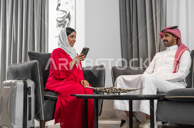 السفر والتنقل لتبادل الخبرات بين الشركات، عقد اجتماعات عمل في الفنادق الراقية، جودة الحياة في الشركات السعودية، رجل وسيدة عربيان خليجيان سعوديان يقومان بالدردشة والتحدث في لوبي الفندق، تحقيق الأهداف وتطوير الخطط، امرأة سعودية ترتدي العباءة والحجاب تستخدم الجوال