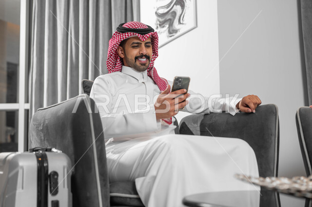 أسلوب الحياة في المملكة العربية السعودية، الجلوس في إحدى الفنادق الراقية، استخدام الجوال والأجهزة التكنولوجية، رجل عربي خليجي سعودي يرتدي الشماغ والثوب التقليدي يجلس في لوبي فندق بجانبه حقيبة السفر ويستخدم الهاتف المحمول
