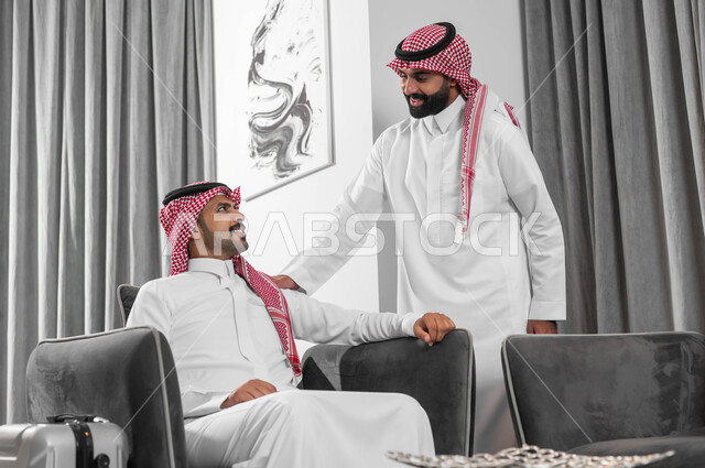 Quality of life in Saudi Arabia, saying hello and greetings with cheerful gestures, holding business meetings in upscale hotels to exchange experiences between the team, two Saudi Gulf Arab men wearing the shemagh and traditional thobe in a hotel lobby, a man greeting his colleague with expressions of pleasure.
