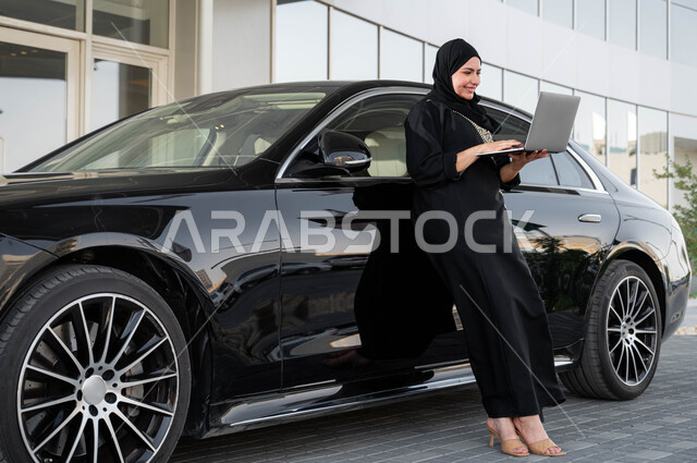 استخدام الاجهزة التقنية، سيده أعمال عربية خليجية سعودية مبتسمة ترتدي الحجاب والعباءة تستند على سيارتها تقوم بالعمل على الحاسوب المحمول, مفهوم الاستقلال والحرية، حرية القيادة للمرأة العربية، القيادة للذهاب الى العمل، اعتماد المرأه على نفسها في التنقل والمواصلات