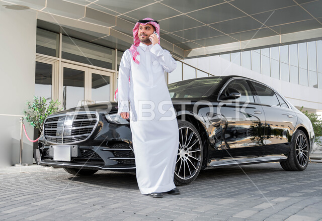 Preparing to drive a car, comfortable quality of life in Saudi Arabia, using advanced technological means, a Saudi Arabian Gulf businessman wearing a shemagh and traditional thobe, leaning on his car, making a phone call via mobile phone