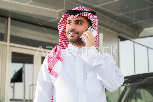 مفهوم الأناقة والاهتمام بالمظهر الخارجي، نمط الحياة المريحة في السعودية، الاستعداد لقيادة السيارة، استخدام الوسائل التقنية المتطورة، صورة مقربة لرجل أعمال عربي خليجي سعودي يرتدي الشماغ والثوب التقليدي يقف أمام سيارته يقوم بإجراء مكالمة هاتفية عبر الهاتف الجوال