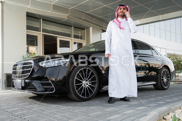 نمط الحياة المريحة في السعودية، الاستعداد لقيادة السيارة، استخدام الوسائل التقنية المتطورة، رجل أعمال عربي خليجي سعودي يرتدي الشماغ والثوب التقليدي يقف أمام سيارته يقوم بإجراء مكالمة هاتفية عبر الهاتف الجوال