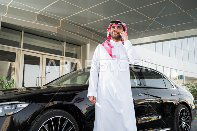 نمط الحياة المريحة في السعودية، الاستعداد لقيادة السيارة، استخدام الوسائل التقنية المتطورة، رجل أعمال عربي خليجي سعودي يرتدي الشماغ والثوب التقليدي يقف أمام سيارته يقوم بإجراء مكالمة هاتفية عبر الهاتف الجوال
