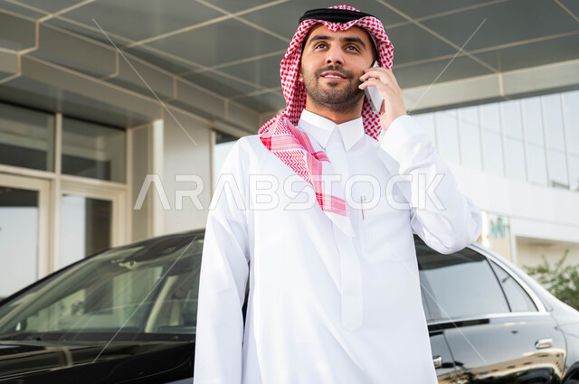 The concept of elegance and attention to external appearance, the comfortable lifestyle in Saudi Arabia, preparing to drive a car, using advanced technical means, a close-up picture of a Saudi Arabian Gulf businessman wearing a shemagh and traditional thobe, standing in front of his car, making a phone call via mobile phone