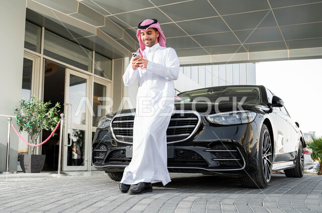 استخدام الوسائل التقنية الحدينة والمتطورة، رجل أعمال عربي خليجي سعودي يرتدي الشماغ والثوب التقليدي يقف أمام سيارته الفاخرة ويحمل بيده الجهاز المحمول، الاستعداد لركوب السيارة، أسلوب الحياة المريح في المملكة العربية السعودية