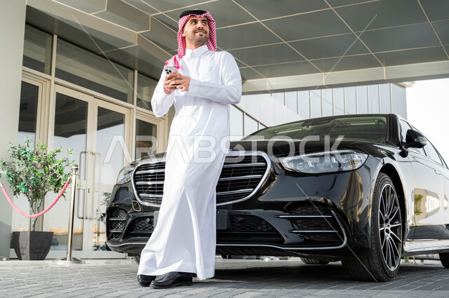 النظر إلى شيء ما، استخدام الوسائل التقنية الحديثة والمتطورة، رجل أعمال عربي خليجي سعودي يرتدي الشماغ والثوب التقليدي يقف أمام سيارته الفاخرة ويحمل بيده الجهاز المحمول، الاستعداد لركوب السيارة، أسلوب الحياة المريح في المملكة العربية السعودية