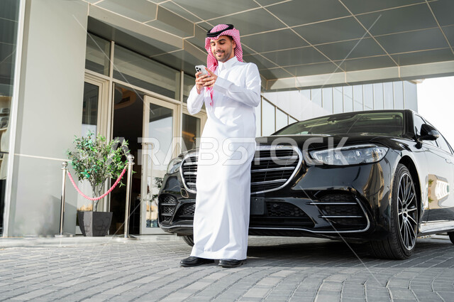 استخدام الوسائل التقنية الحدينة والمتطورة، رجل أعمال عربي خليجي سعودي يرتدي الشماغ والثوب التقليدي يقف أمام سيارته الفاخرة ويحمل بيده الجهاز المحمول، الاستعداد لركوب السيارة، أسلوب الحياة المريح في المملكة العربية السعودية
