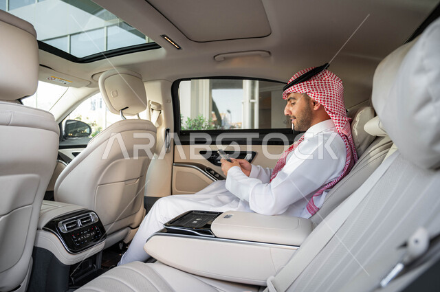 خدمات النقل الآمنة والمريحة في المملكة، خدمة توصيل الركاب، استخدام الوسائل التقنية الحديثة والمتطورة، رجل أعمال عربي خليجي سعودي يرتدي الشماغ والثوب التقليدي يجلس داخل سيارة الفاخرة ويستخدم الهاتف المحمول، أسلوب الحياة المريح في السعودية