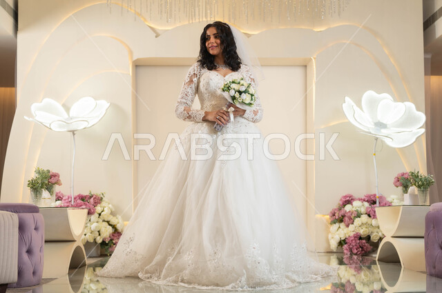قاعات الأفراح والمناسبات المزينة بالزهور، مراسم الأعراس والاحتفالات في المملكة، عروس عربية خليجية سعودية جميلة ترتدي فستان الزفاف الأبيض تمسك بيدها باقة ورد، الفرح و السرور في يوم الزواج، الاهتمام بالأناقة و المظهر الخارجي، مفهوم التزين والاطلالة المتألقة