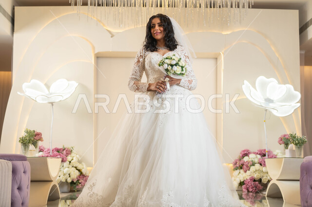 الاهتمام بالأناقة و المظهر الخارجي، مفهوم التزين والاطلالة المتألقة، قاعات الأفراح والمناسبات المزينة بالزهور، مراسم الأعراس والاحتفالات في المملكة، عروس عربية خليجية سعودية جميلة ترتدي فستان الزفاف الأبيض تمسك بيدها باقة ورد، الفرح و السرور في يوم الزواج