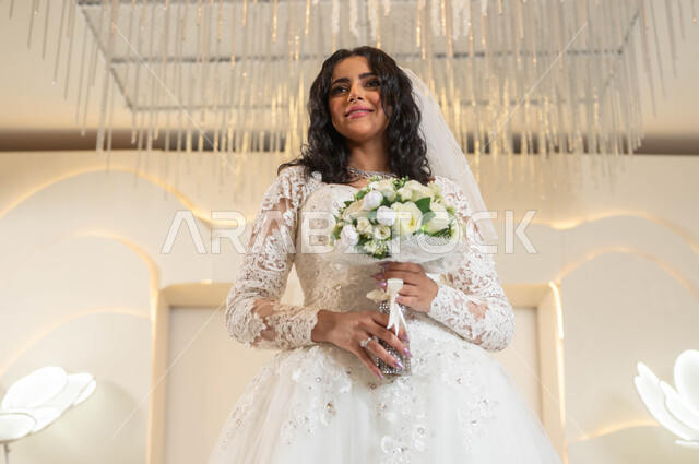 الفرح والسرور في يوم الزواج، مفهوم التأنق والتجمل، صورة مقربة لعروس عربية خليجية سعودية جميلة ترتدي فستان الزفاف الأبيض تمسك بيدها باقة ورد، الاهتمام بالأناقة و المظهر الخارجي، التزين والاطلالة المتألقة، قاعات الأفراح والمناسبات المزينة بالزهور