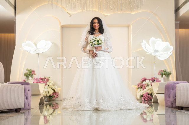 الوقوف باستقامة والنظرإلى الكاميرا بسعادة، عروس عربية خليجية سعودية جميلة ترتدي فستان الزفاف الأبيض تمسك بيدها باقة ورد، الفرح و السرور في يوم الزواج، الاهتمام بالأناقة و المظهر الخارجي، مفهوم التزين والاطلالة المتألقة، قاعات الأفراح والمناسبات المزينة بالزهور