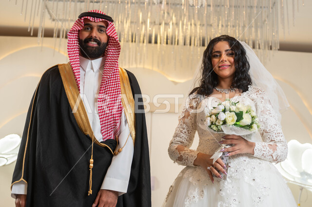 الأناقة والاهتمام بالمظهر الخارجي, صورة لعريس و عروس عربيان خليجيان سعوديان في قاعة الزفاف ينظران إلى الكاميرا بإيماءات السعادة، المناسبات والأفراح الخاصة, مفهوم الاستقرار وتكوين الأسرة, السرور والفرح في يوم الزواج، مراسم الأعراس في المملكة