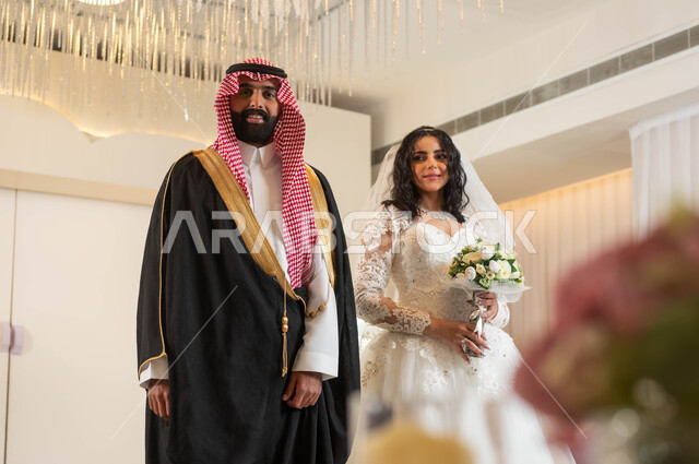مراسم الاعراس و المناسبات في المملكة، الفرح و السرور في يوم الزفاف، اجمل إطلالة لرجل عربي وسيم يرتدي الزي الرسمي والبشت التقليدي، صورة لعروسة عربية خليجية سعودية ترتدي فستان زفاف أبيض تمسك بيدها باقة ورد، مفهوم الاستقرار وتكوين عائلة، أصالة العادات والتقاليد