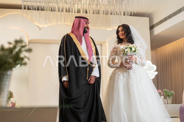 السرور والفرح في يوم الزواج، تبادل نظرات الحب، عروس عربية خليجية سعودية جميلة تمسك بيدها باقة ورود، عريس عربي وسيم يقف بهيبة و رجولة، الأناقة والاهتمام بالمظهر الخارجي, المناسبات والأفراح الخاصة, مفهوم الاستقرار وتكوين الأسرة, مراسم الأعراس في المملكة