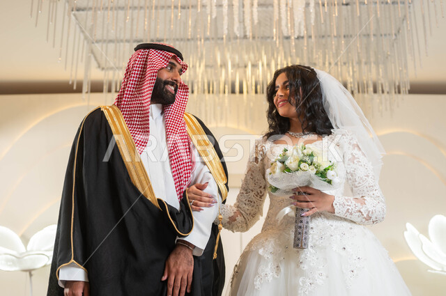 تبادل نظرات الحب، عروس عربية خليجية سعودية جميلة تمسك بيدها باقة ورود، عريس عربي وسيم يقف بهيبة و رجولة، الأناقة والاهتمام بالمظهر الخارجي, المناسبات والأفراح الخاصة, مفهوم الاستقرار وتكوين الأسرة, السرور والفرح في يوم الزواج، مراسم الأعراس في المملكة