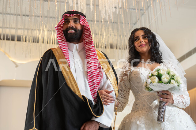 The most beautiful view of a handsome Arab man wearing a formal uniform and a traditional bisht, the authenticity of customs and traditions, wedding ceremonies and events in the Kingdom, joy and pleasure on the wedding day, a picture of a Saudi Gulf Arab bride wearing a white wedding dress holding a bouquet of roses in her hand, the concept of stability and forming a family