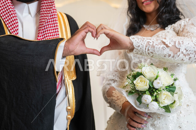 الحب و المودة بين الأزواج، إيماءات يدين على شكل قلب، عروس عربية خليجية سعودية جميلة ترتدي فستان زفاف أبيض، صورة مقربة لعريس عربي وسيم، مراسم الأعراس و المناسبات في المملكة، السرور والفرح في يوم الزواج، مفهوم الوفاء بالوعود، قاعات الافراح المزينة