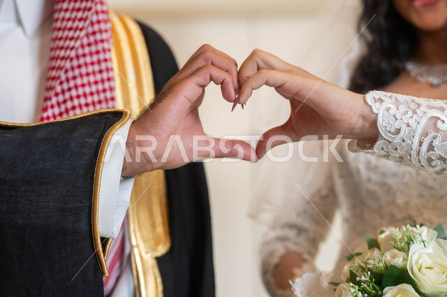 إيماءات يدين على شكل قلب، الحب و المودة بين الأزواج، عروس عربية خليجية سعودية جميلة ترتدي فستان زفاف أبيض، صورة مقربة لعريس عربي وسيم، مراسم الأعراس و المناسبات في المملكة، السرور والفرح في يوم الزواج، مفهوم الوفاء بالوعود، قاعات الافراح المزينة