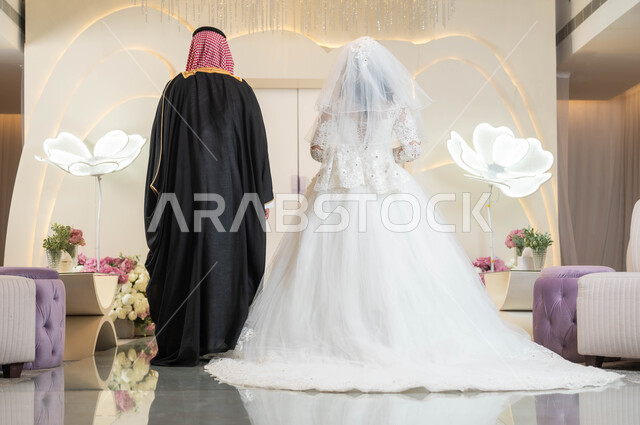 الاستقرار وتكوين عائلة، عروس عربية جميلة تمسك بيد زوجها، مفهوم الوفاء بالوعود، مراسم الأعراس والمناسبات في المملكة، قاعات الأفراح المزينة بالزهور، صورة من الخلف لعريس عربي خليجي سعودي، السرور والفرح في يوم الزواج، الاهتمام بالمظهر الخارجي