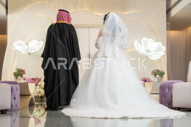 عروس عربية جميلة تمسك بيد زوجها، مفهوم الوفاء بالوعود، مراسم الأعراس والمناسبات في المملكة، قاعات الأفراح المزينة بالزهور، صورة من الخلف لعريس عربي خليجي سعودي، الاستقرار وتكوين عائلة، السرور والفرح في يوم الزواج، الاهتمام بالمظهر الخارجي