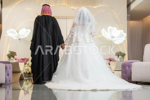 The concept of fulfilling promises, wedding and event ceremonies in the Kingdom, wedding halls decorated with flowers, a beautiful Arab bride holding her husband’s hand, a picture from behind of a Saudi Gulf Arab groom, stability and forming a family, happiness and joy on the wedding day, attention to external appearance