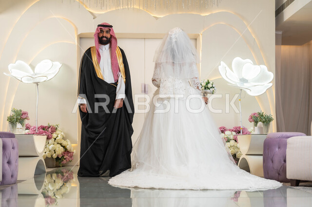 عريس يرتدي الزي الرسمي والبشت التقليدي يقف مع عروسه بشكل متعاكس، قاعات الأفراح المزينة بالأزهار، الاناقة والاهتمام بالمظهر الخارجي، عروسة عربية خليجية سعودية جميلة ترتدي فستان زفاف أبيض، مراسم الاعراس والمناسبات في المملكة، الفرح والسرور في يوم الزواج