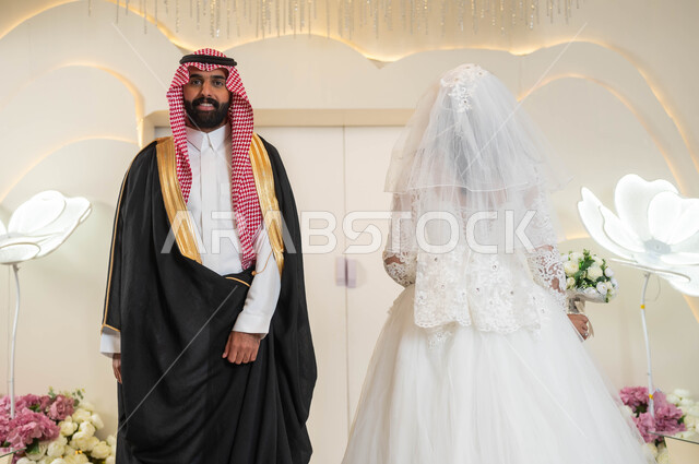 الاناقة والاهتمام بالمظهر الخارجي، عريس يرتدي الزي الرسمي والبشت التقليدي يقف مع عروسه بشكل متعاكس، عروسة عربية خليجية سعودية جميلة ترتدي فستان زفاف أبيض، مراسم الاعراس والمناسبات في المملكة، الفرح والسرور في يوم الزواج، قاعات الأفراح المزينة بالأزهار