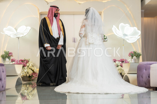 قاعات الأفراح المزينة بالأزهار، الاناقة والاهتمام بالمظهر الخارجي، عريس يرتدي الزي الرسمي والبشت التقليدي يقف مع عروسه بشكل متعاكس، عروسة عربية خليجية سعودية جميلة ترتدي فستان زفاف أبيض، مراسم الاعراس والمناسبات في المملكة، الفرح والسرور في يوم الزواج