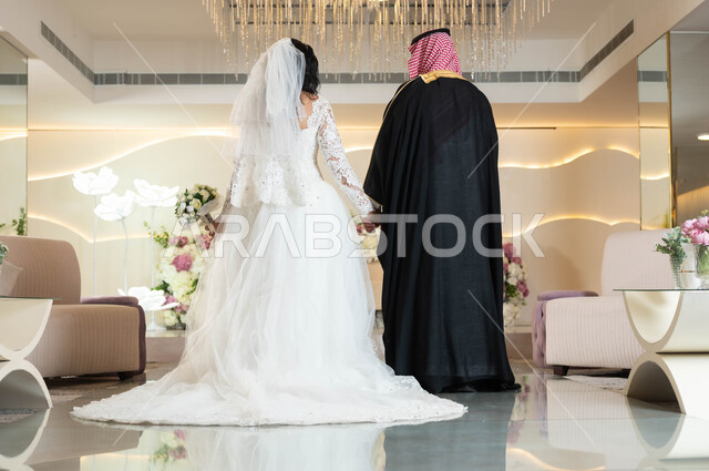 قاعات الأفراح المزينة بالورود و الزهور، عروس عربية جميلة تمسك بيدها باقة ورد، صورة من الخلف لعريس عربي خليجي سعودي يمسك بيد عروسه، الاستقرار وتكوين عائلة، مفهوم الوفاء بالوعود، السرور والفرح في يوم الزواج، مراسم الأعراس والمناسبات في المملكة، صورة لكامل الجسم