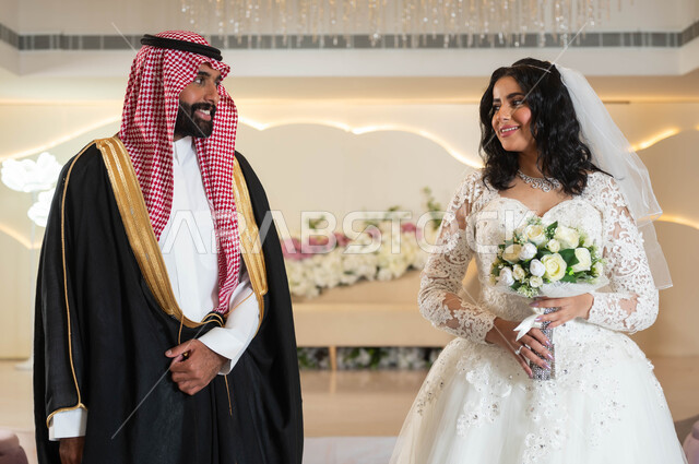 تبادل نظرات الحب، عروس عربية خليجية سعودية جميلة تمسك بيدها باقة ورود، عريس عربي وسيم يقف بهيبة و رجولة، الأناقة والاهتمام بالمظهر الخارجي, المناسبات والأفراح الخاصة, مفهوم الاستقرار وتكوين الأسرة, السرور والفرح في يوم الزواج، مراسم الأعراس في المملكة