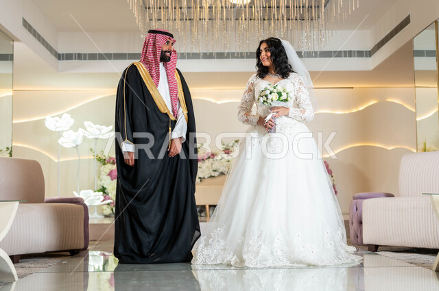 تبادل نظرات المودة والحب، السرور والفرح في يوم الزواج، عروس عربية خليجية سعودية جميلة تمسك بيدها باقة ورود، عريس عربي وسيم يقف بهيبة و رجولة، الأناقة والاهتمام بالمظهر الخارجي, المناسبات والأفراح الخاصة, مفهوم الاستقرار وتكوين الأسرة, مراسم الأعراس في المملكة